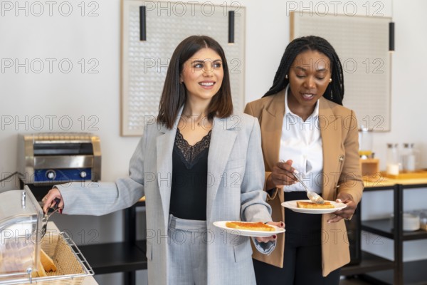 Two businesswomen are enjoying breakfast at a hotel buffet, adding jam to their toast during a business trip