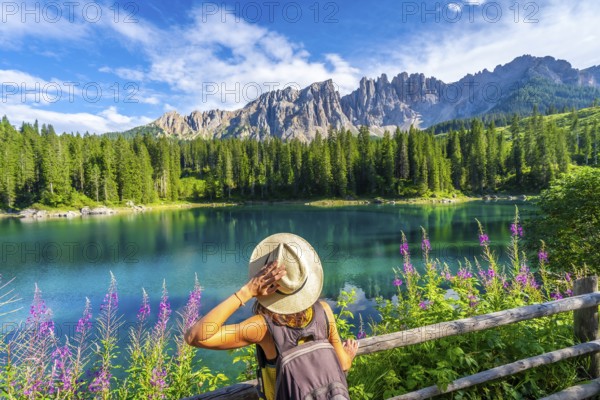 Woman with backpack and hat admiring the turquoise waters of lake carezza with the latemar mountain range in the background, dolomites, italy