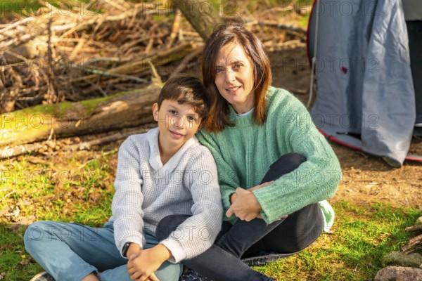 Caucasian mother and son smiling at camera sunbathing enjoying nature in the forest next to camping tent