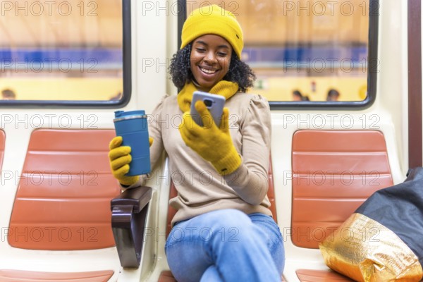Smiling black woman wearing yellow winter clothes using phone and drinking coffee while traveling by subway train