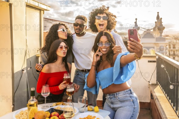 Happy multi-ethnic friends taking selfie celebrating evening party in rooftop drinking wine and eating snacks