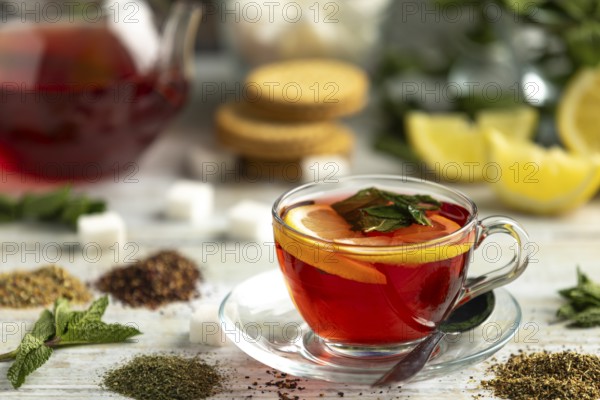 A cup of red tea with lemon and mint surrounded by sugar cubes, teas and biscuits