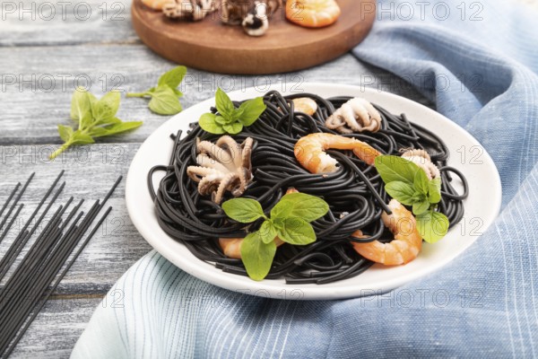 Black cuttlefish ink pasta with shrimps or prawns and small octopuses on gray wooden background and blue textile. Side view, close up