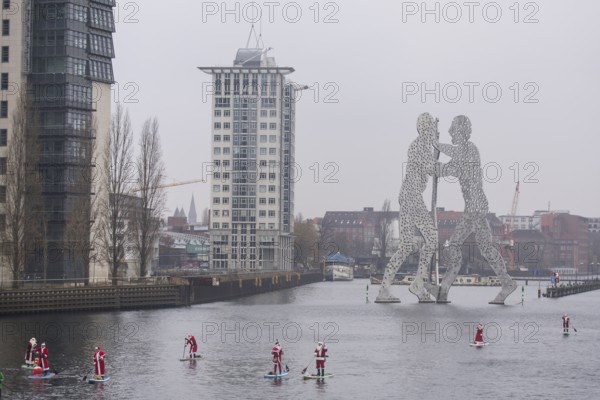 Water sports enthusiasts dressed as Santas ride SUPs on the Spree in front of the Molecule Man in Berlin on 14 December 2024