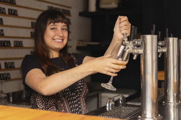 A cheerful waitress serves a glass of beer from a tap in a modern bar. She smiles and enjoys her work