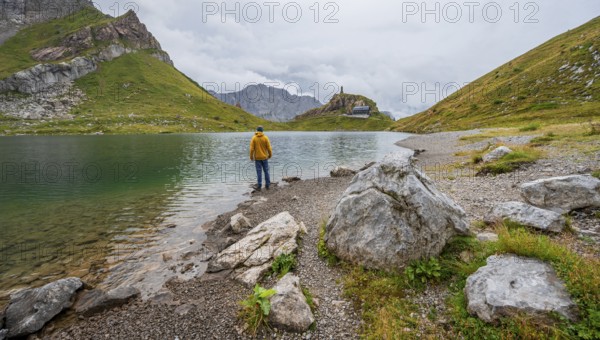 Mountaineer in yellow jacket at the mountain lake, view over the Wolayersee lake to the alpine club hut Wolayerseehütte, cloudy mountain landscape, Carnic Alps, Carnic High Trail, Carinthia, Austria, Europe