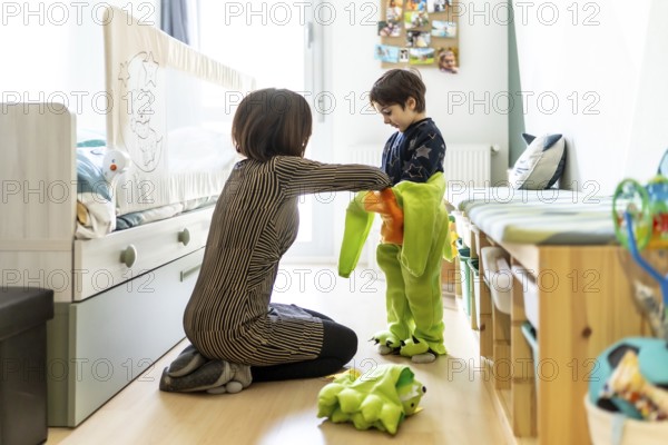 Mother assisting her son in getting dressed in a dinosaur costume, fostering imaginative play in a cozy bedroom setting