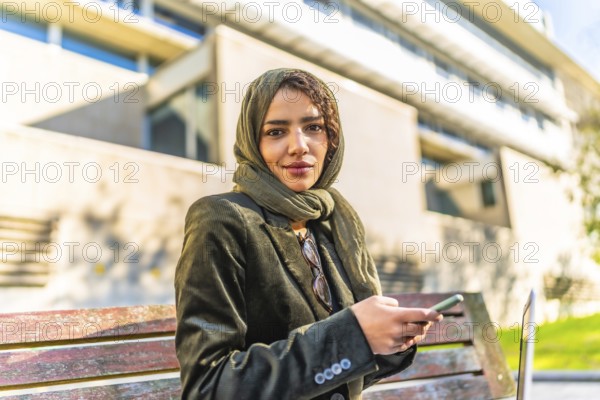 Muslim businesswoman using smartphone sitting on a park bench next to office building