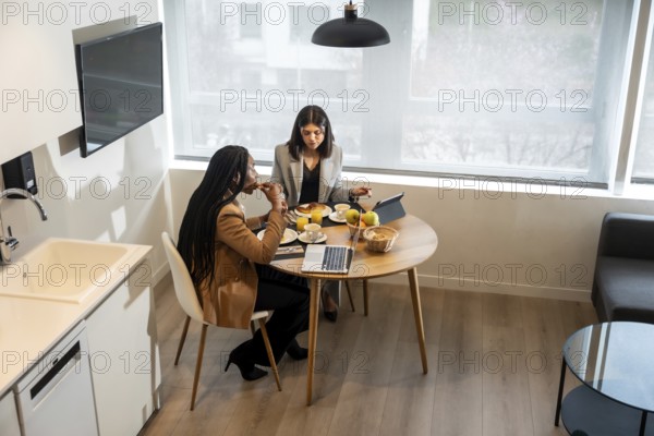Two businesswomen are having breakfast in their hotel room while working on a tablet and laptop during a business trip