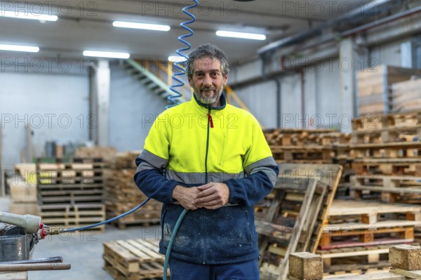 Portrait of a mature man manual worker with reflective clothes in a recycling plant full of pallets to arrange to reuse