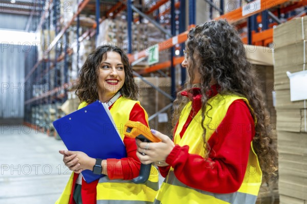 Two young women talking while taking inventory in a distribution warehouse using clipboard and scanner