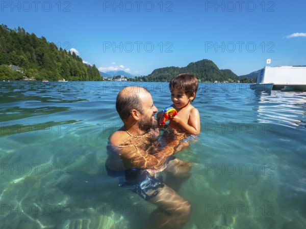 Father playing with his son in the clear water of lake bled, with mountains and forest in the background, enjoying summer vacation