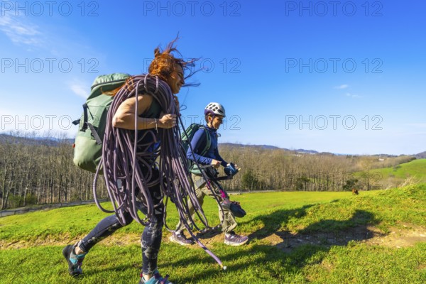 Two female climbers are walking across a mountain meadow carrying climbing rope and gear on a sunny day