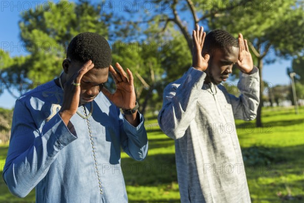 Two young senegalese men wearing traditional dashiki clothing are praying outdoors in a park, observing ramadan