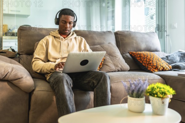 Young man concentrating on his laptop while wearing headphones, comfortably seated on a sofa in his modern living room, working or studying from home