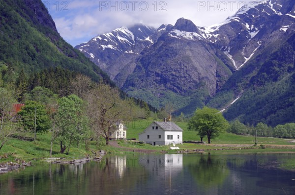 A white house on a calm lake, surrounded by green trees and majestic snow-capped mountains under a cloudy sky, Gaular, Balestrand, Norway, Europe