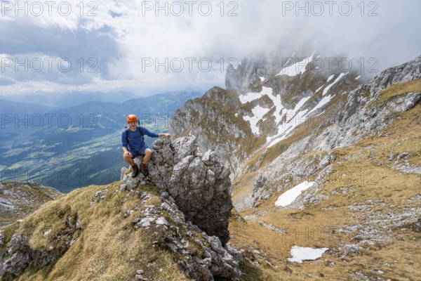 Mountaineer with helmet sitting on a rock, ascent to the Maukspitze, clouds moving around the mountains, Wilder Kaiser, Kaiser Mountains, Tyrol, Austria, Europe