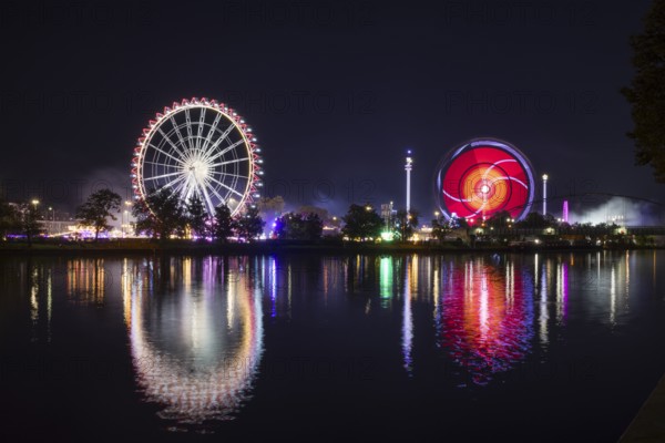 Atmospheric funfair at night with brightly lit Ferris wheel and colourful lights reflected in the water, Cannstadter Volksfest, Stuttgart, Baden-Württemberg