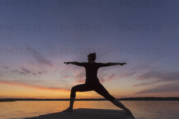 Woman practicing yoga posture Virabhadrasana II, Warrior Pose, lunging standing asana on jetty at lake at sunset