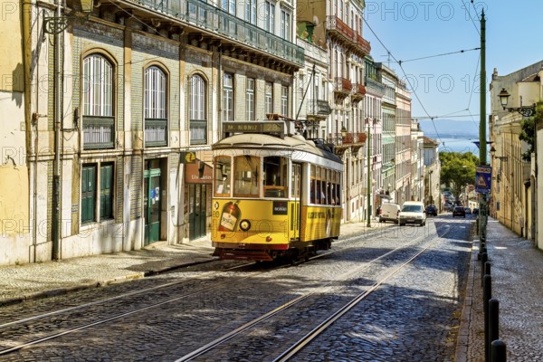 A historic yellow tram travels down a sunny Lisbon street lined with old buildings under a clear blue sky, Alfama, Lisbon