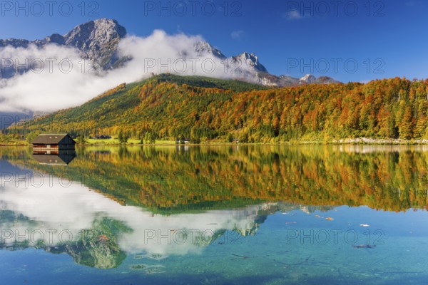 Lake Almsee in autumn with a forest reflected in the lake. A slowly dissipating fog moves over the lake. Blue sky with no clouds