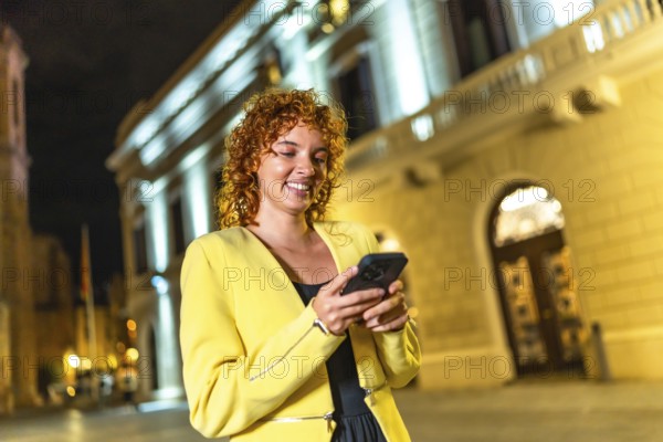 Smiling cheerful woman in yellow clothes and curly hair using smartphone walking along city at night