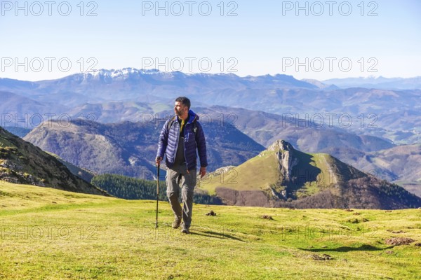 Male hiker walking on grassy mountain slope with trekking poles, enjoying scenic view of mount txindoki and surrounding peaks in gipuzkoa, basque country, spain