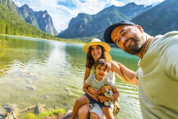 Happy family taking a selfie by the transparent water of lake dobbiaco with the dolomites in the background, enjoying summer vacation