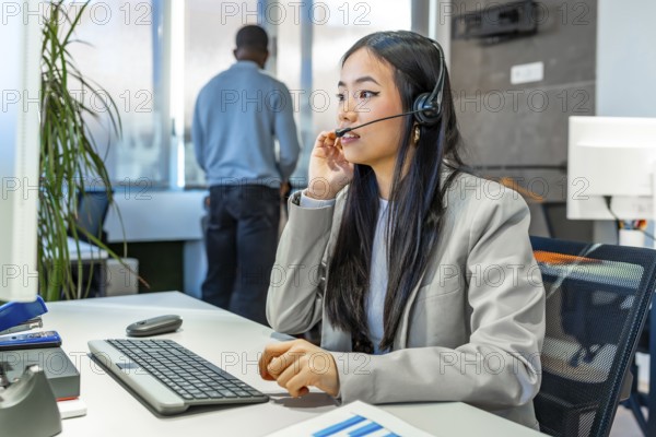 Young asian manager working diligently at a computer, wearing a headset in a sleek, modern coworking office environment
