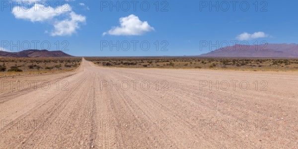 An endless desert road under a blue sky with a wide, dry landscape and mountains in the background, gravel road D2315, Namibia, Africa