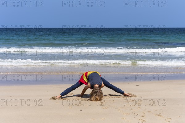A woman doing yoga on a sandy beach by the sea. Prasarita Padottasana or forward bend with legs apart. Treen Cove, Cornwall, England, Great Britain