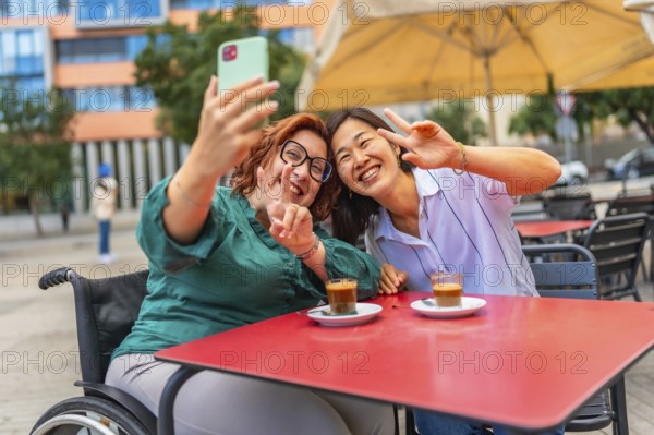 Cheerful disabled woman and asian female friend gesturing success taking selfie sitting in an outdoor cafeteria in the city