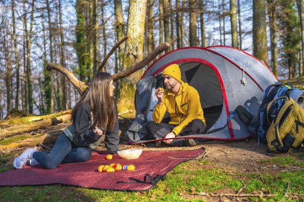 Couple of hikers having a snack next to their tent during a camping trip in the forest, enjoying the sunny weather