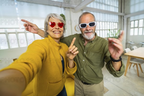 Frontal personal point of view of a happy senior couple with fun sunglasses taking selfie at apartment