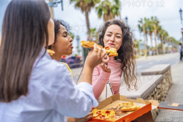 Three female diverse tourists chatting and sharing pizza sitting in the street