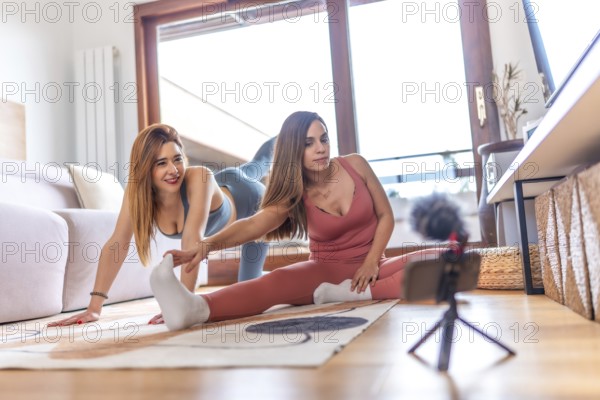 Two young women recording an online yoga class at home, performing stretching exercises in the living room