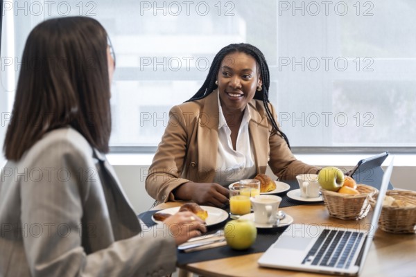 Two businesswomen enjoying breakfast and engaging in conversation during their business trip, utilizing laptop and tablet