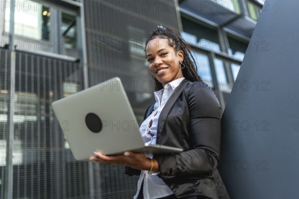 Modern latin businesswoman with long braids working using laptop standing outside a financial building