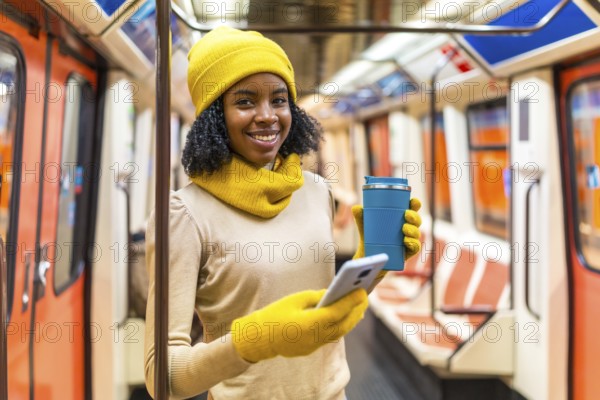 Smiling woman wearing yellow winter clothes using smartphone and drinking coffee inside subway train