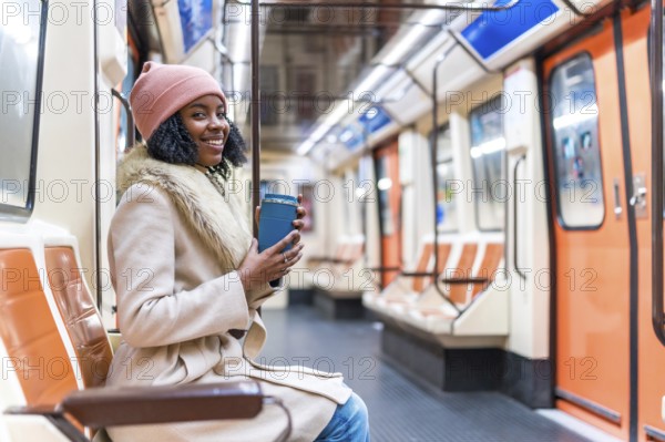 Smiling young black woman wearing winter clothes holding reusable coffee cup while riding subway train