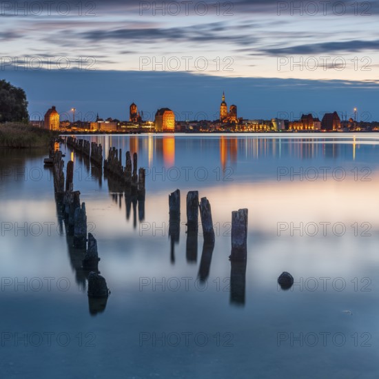 View at dusk over the Strelasund to the illuminated town of Stralsund with its churches and the warehouses in the harbour, old groynes in front, Stralsund, Mecklenburg-Western Pomerania, Germany, Europe