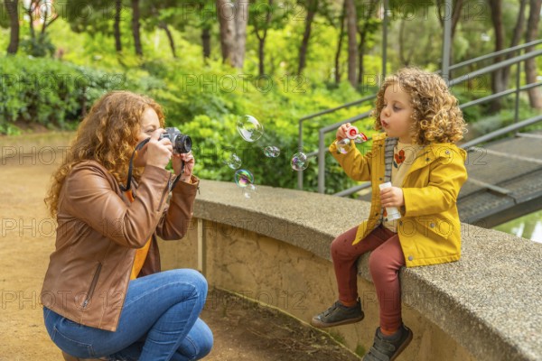 Caucasian adult woman taking a photo with a digital camera of her daughter blowing soap bubbles in a park