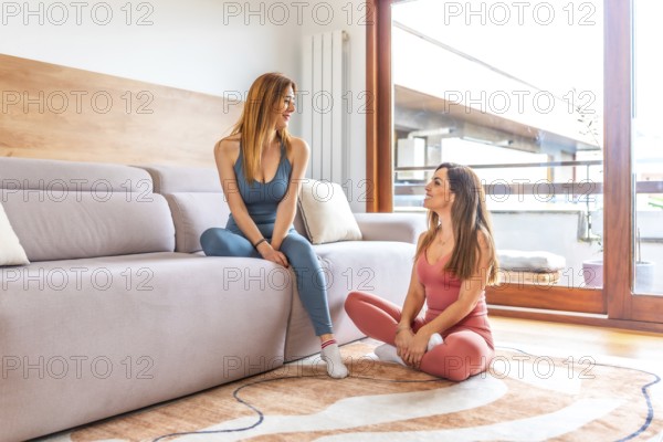 Two sporty women taking a break on a sofa and a carpet in the living room, resting after an online yoga class at home
