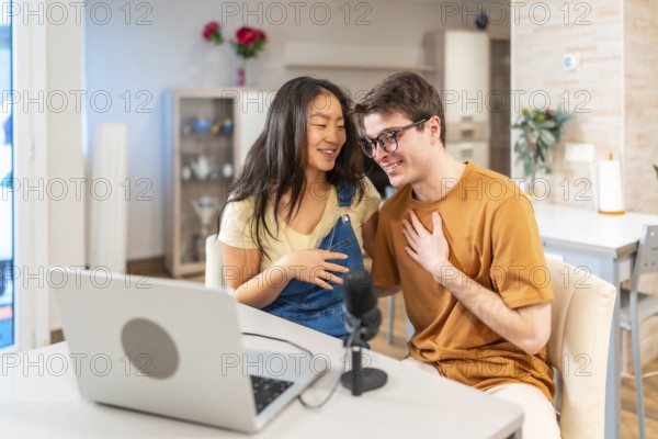 Young multiethnic couple sitting together at home, using a laptop and microphone to record their engaging audio podcast