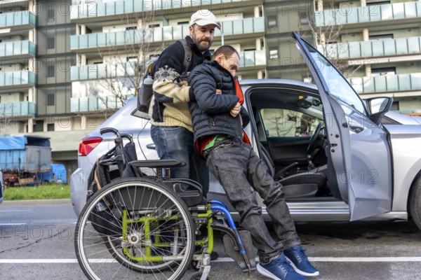 Caregiver lifting a disabled young man from his wheelchair and helping him to get into a car parked in the city