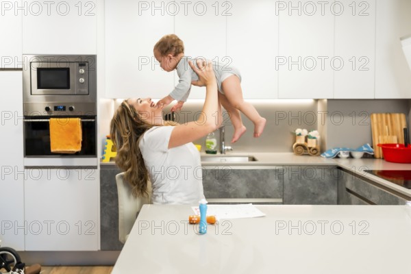 Happy mother playing with baby, lifting her in the air in a modern kitchen, enjoying time together