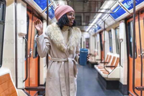 Smiling young black woman wearing winter coat and pink woolen hat holding reusable coffee cup waving hand inside subway train