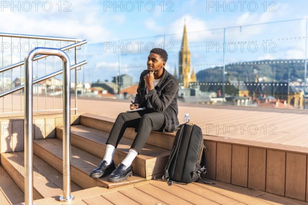 Pensive african young male traveler writing notes sitting on a city viewpoint during sunny day