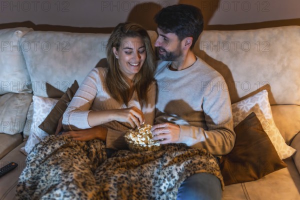Romantic couple sharing a bowl of popcorn while watching a movie on the sofa at night, enjoying a cozy and intimate moment together