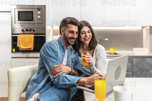 Young couple laughing while watching a movie on a laptop in the kitchen and drinking orange juice